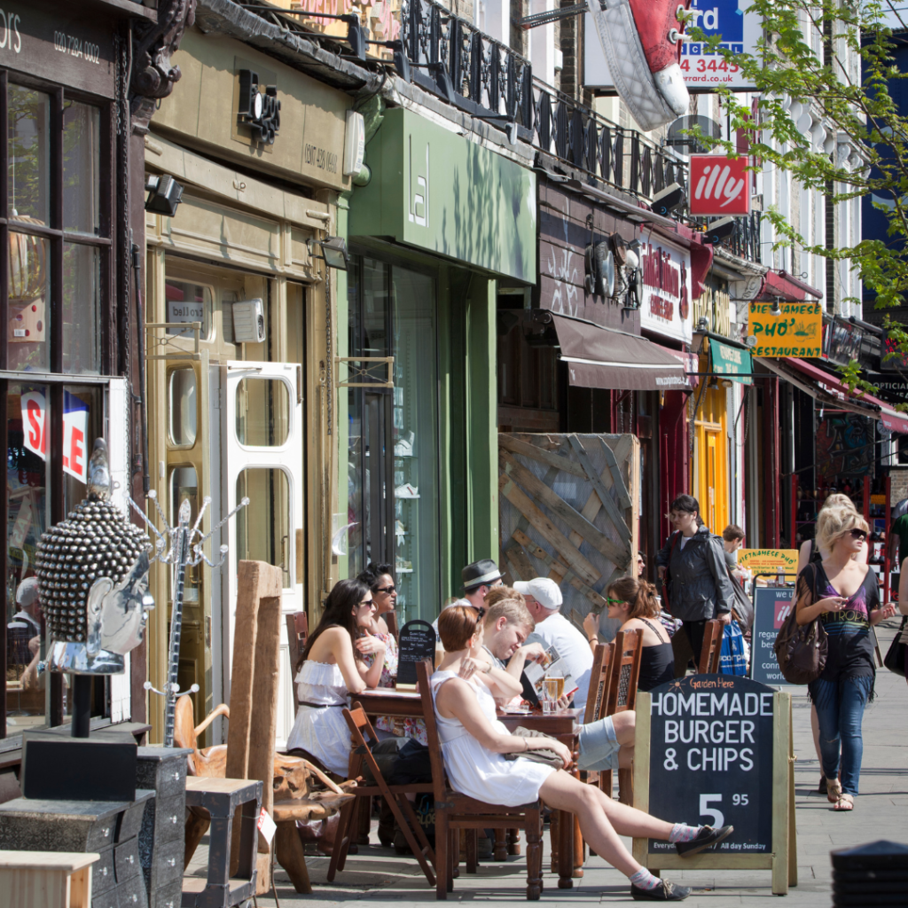 A row of shops in Camden, London with a number of people sitting outside a cafe. A sign reads "Homemade burger and chips"